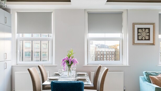 A dining area with a table set for four, two windows with blinds, a radiator, and a framed picture on the wall. - Stylish Modern 2-bedroom Apartment on Cromwell (London)