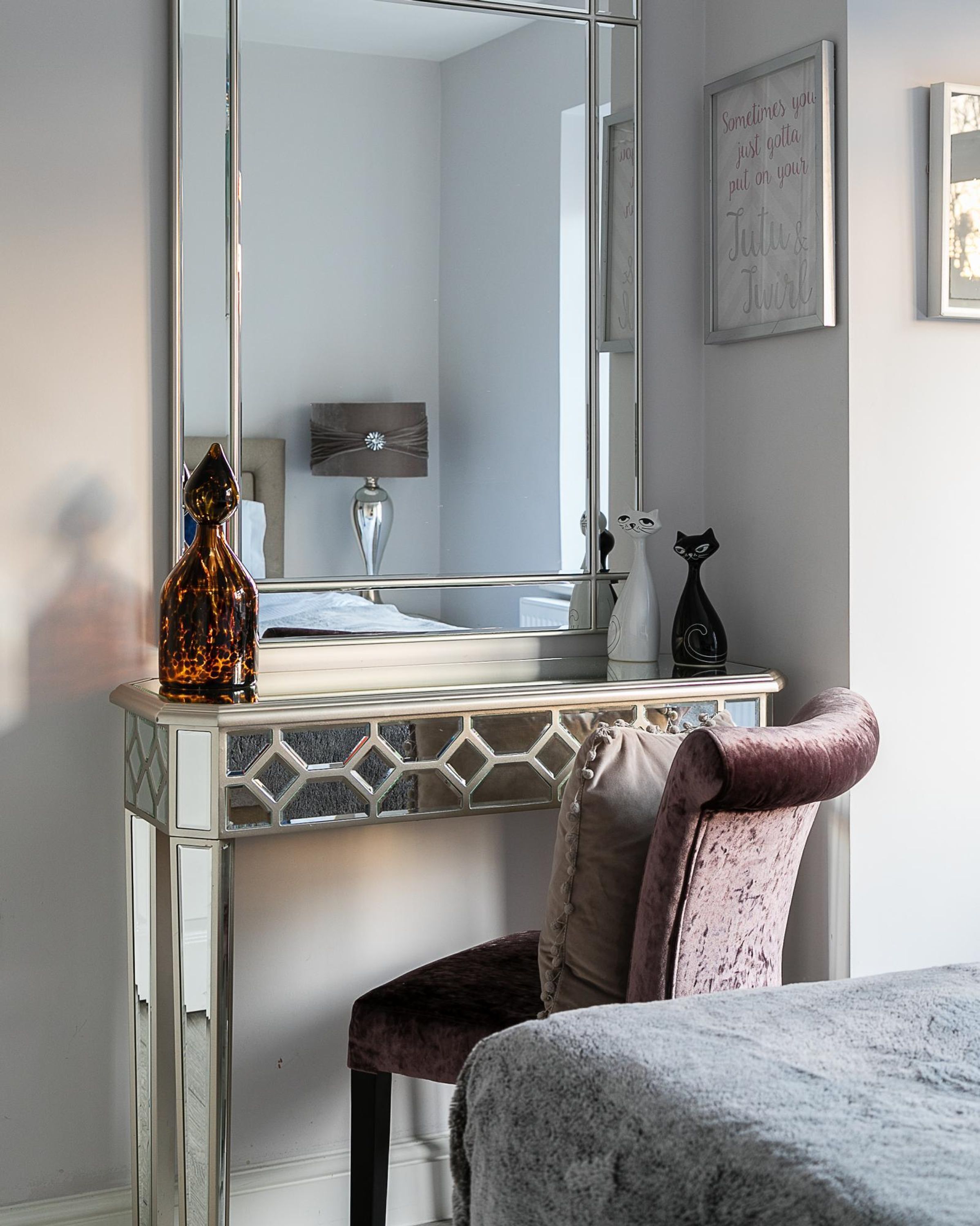 Une chambre à coucher comprenant une coiffeuse, un miroir, un vase décoratif, une statuette de chat noir et un lit recouvert d’une couverture à motifs.