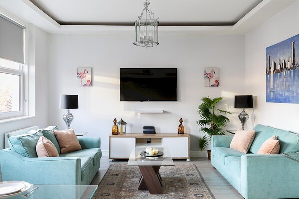 A modern living room with two teal sofas, a glass coffee table, a flat-screen TV, and decorative items. - Stylish Modern 2-bedroom Apartment on Cromwell (London)