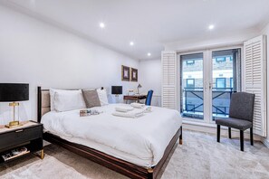 A modern bedroom with a large bed, bedside table, lamp, and a view of the outside through a window with shutters. - At Home in Marylebone (London)