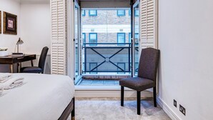 A bedroom with a bed, a chair, a desk, and a window with a view of buildings. - At Home in Marylebone (London)
