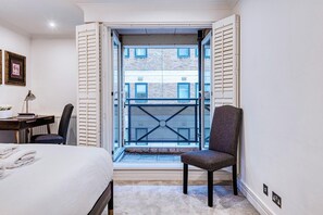 A bedroom with a bed, a chair, a desk, and a window with a view of buildings. - At Home in Marylebone (London)