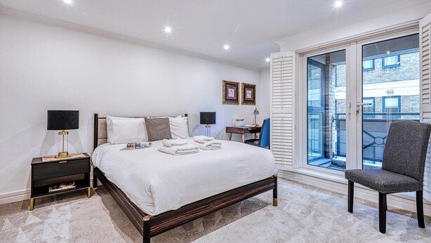 A modern bedroom with a large bed, bedside tables, a chair, and a view of the outside. - At Home in Marylebone (London)