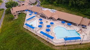 Vista aerea di un resort con un’ampia area piscina, diverse zone relax e un edificio caratterizzato da un design del tetto originale.