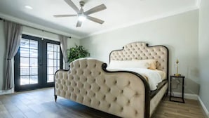A bedroom with a tufted headboard bed, a ceiling fan, and a large window with curtains.