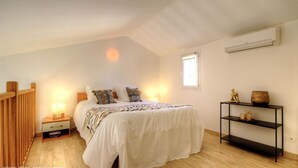 Quarto com cama, abajures, mesa de cabeceira, um banco de madeira, uma pequena mesa com objetos decorativos e uma janela.