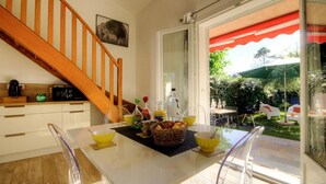 Uma cozinha moderna com escada de madeira, área de refeições com cadeiras amarelas e vista para um jardim com mobiliário de exterior.