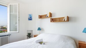Une chambre à coucher bien rangée, avec un grand lit, une fenêtre donnant sur des immeubles, deux étagères murales remplies de livres et une petite plante.