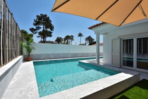 Une piscine rectangulaire à l’eau d’un bleu limpide, entourée d’une terrasse carrelée de couleur blanche. Un grand parasol beige offre de l’ombre, et on aperçoit un pot de fleurs sur la gauche.