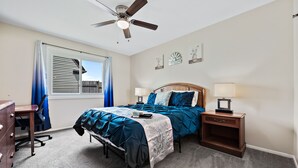 Une chambre à coucher comprenant un lit, un bureau, une chaise, une table de chevet, une lampe, un ventilateur de plafond et une fenêtre avec des rideaux.