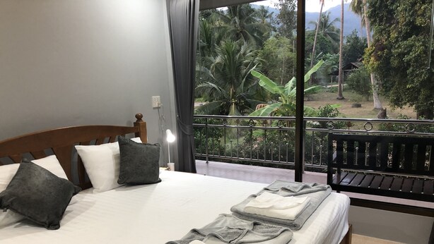 Een hotelkamer met een bed, kussens, een nachtkastje, een lamp en uitzicht op een balkon met groen.