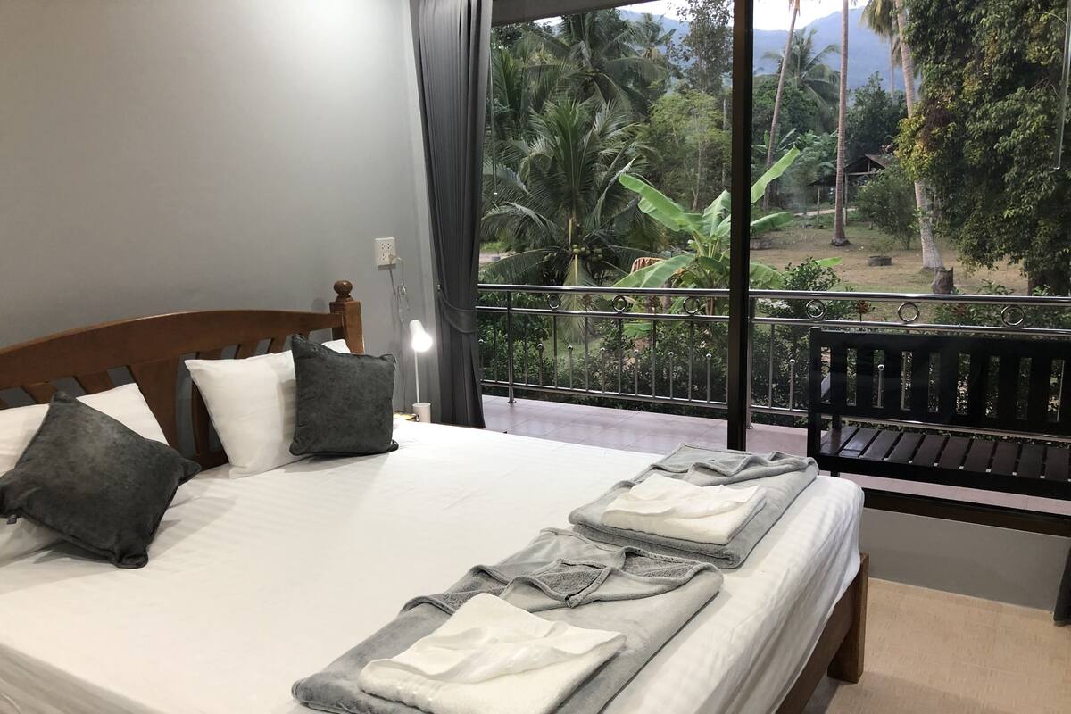 Ein Hotelzimmer mit Bett, Kissen, Nachttisch, Lampe und Blick auf einen Balkon mit Grünpflanzen.