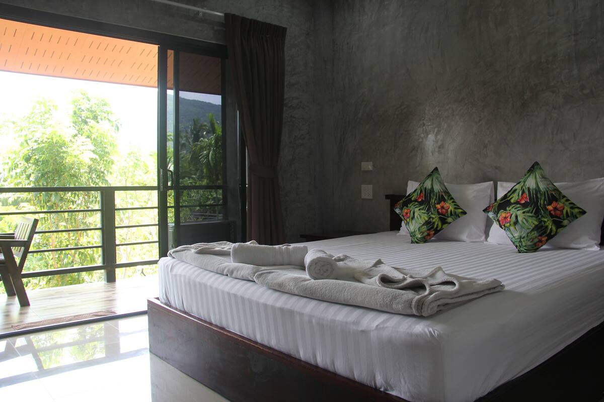 Ein Hotelzimmer mit einem großen Bett, zwei Kissen mit tropischen Motiven, einem Holzstuhl und einem Balkon mit Blick auf Grünflächen.