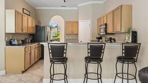 A kitchen with wooden cabinets, a refrigerator, a microwave, a toaster, and a coffee maker. There are three bar stools and a window with a view of greenery.