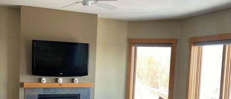 A living room with a fireplace, a television, a ceiling fan, and a large window.