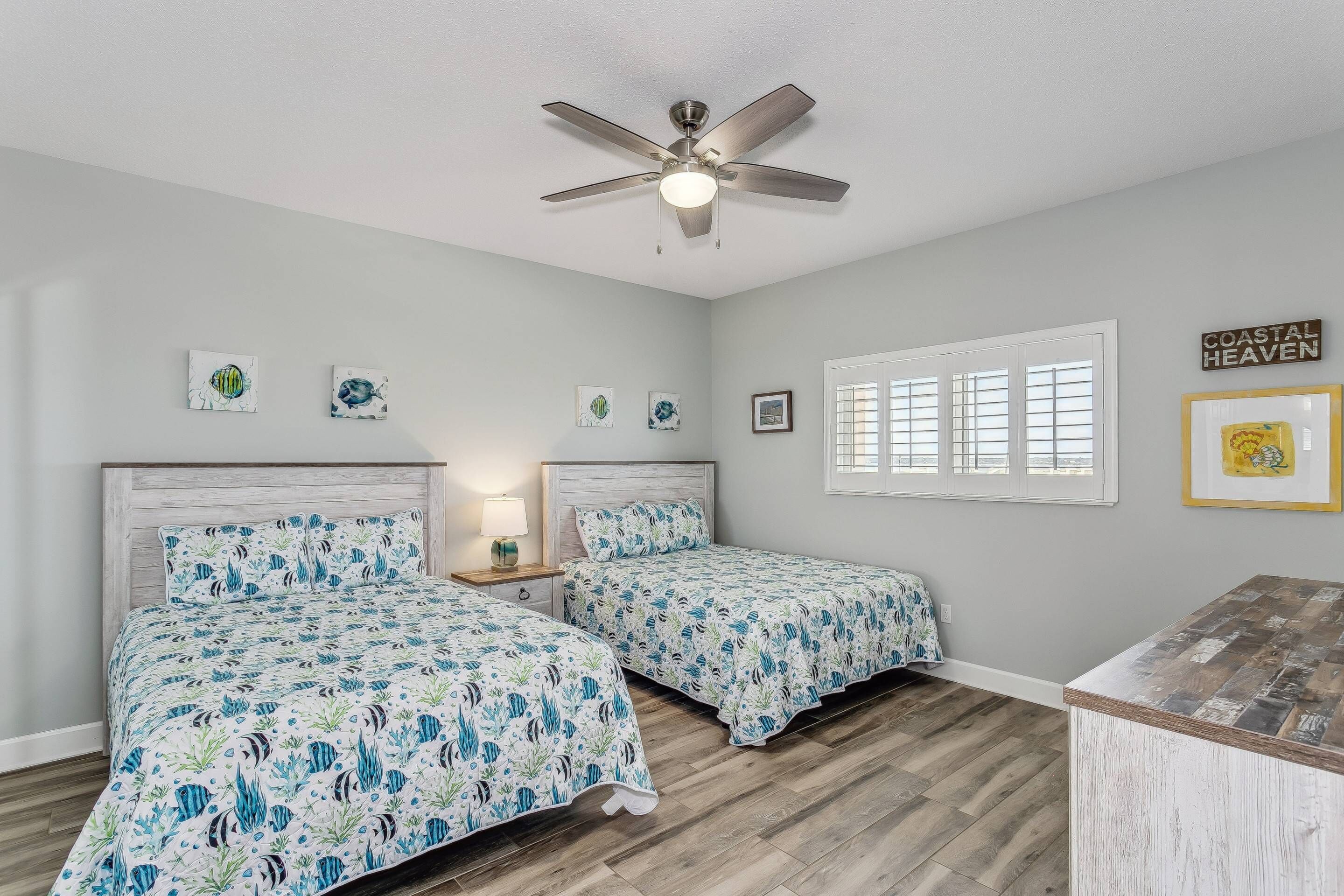 A bedroom with two beds, a ceiling fan, a window with blinds, and artwork on the walls.