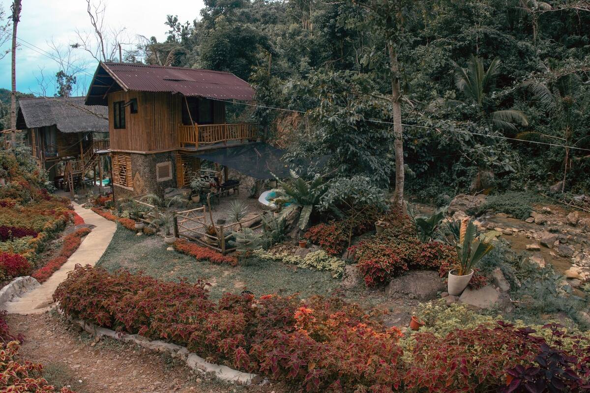 Een rustiek houten huis met een balkon, omgeven door een tuin met verschillende planten en een klein beekje.