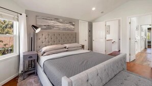 A bedroom with a large bed, a bedside lamp, a grey upholstered bench, and a view of the outdoors. - Pacifica Storybook Beach House, 2 blocks from beach, pier and golf, 10 min to SF (Pacifica)
