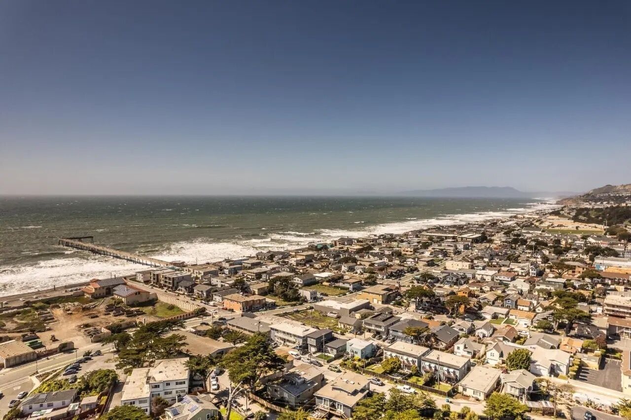 A coastal town with a pier extending into the sea, residential buildings, and a clear view of the ocean.