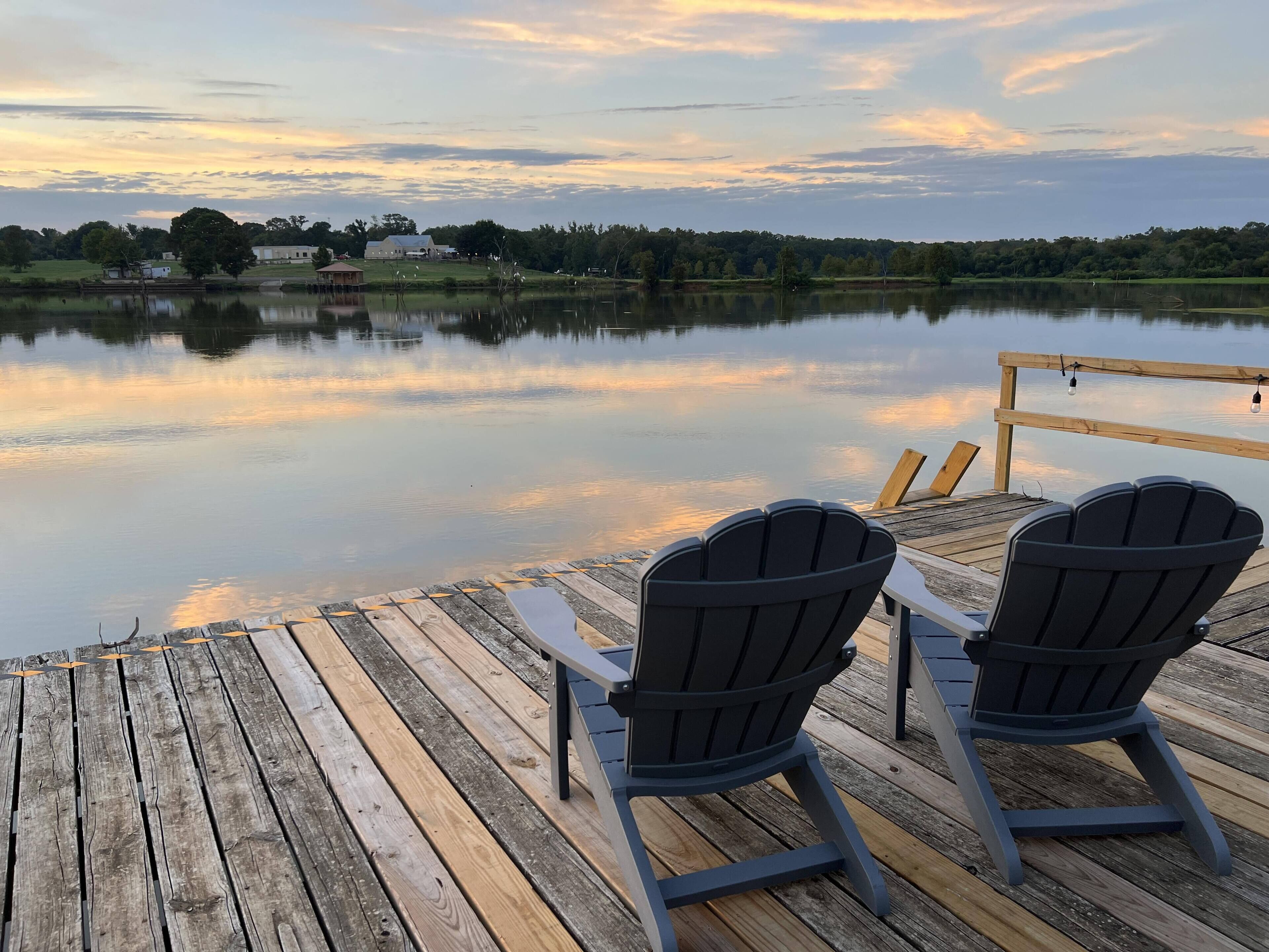 A wooden dock with two Adirondack chairs facing a calm lake at sunset.