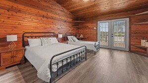 Ein schlicht eingerichtetes Schlafzimmer im Blockhausstil mit zwei Betten, Holzwänden, einem Deckenventilator und einer Schiebetür zum Außenbereich.