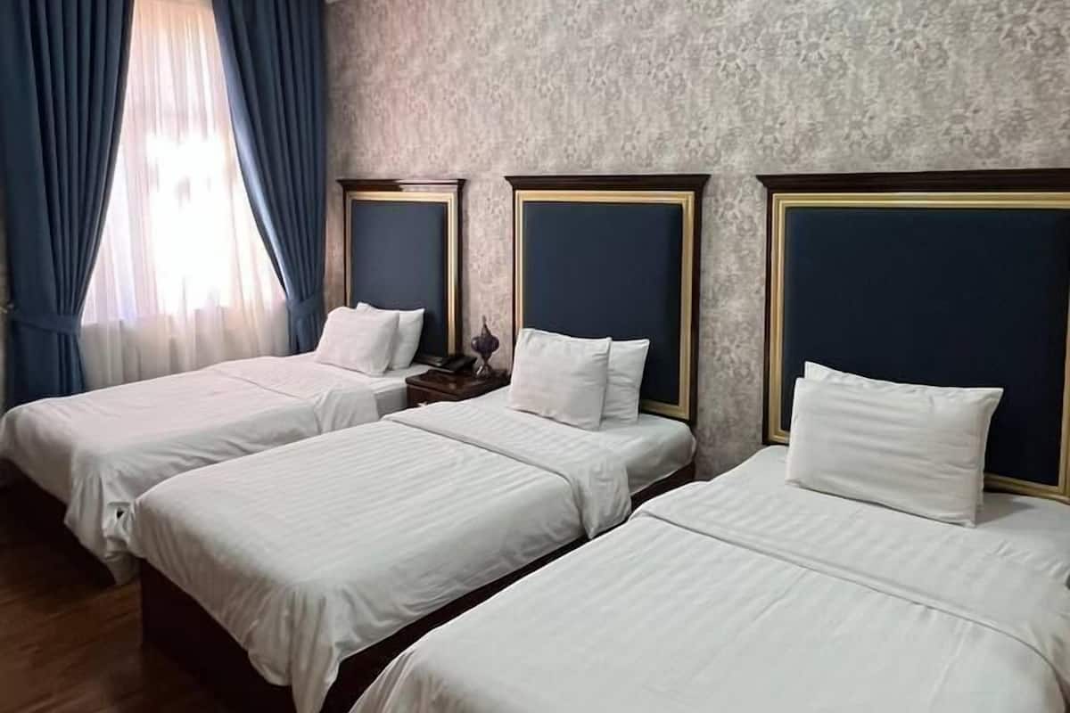 A hotel room with three single beds, wooden flooring, a patterned wall, and a window with blue curtains.