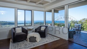 A spacious room with large windows offering a view of the sea and landscape. There are two dark armchairs with white cushions and a small round table.