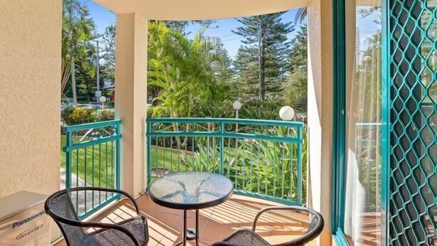 A balcony with a glass table and two chairs, surrounded by a green railing and a view of tropical trees. - Aruba Beach - Private Apartments (Broadbeach)