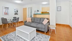 A modern living room with a grey sofa, white coffee table, and a small side table. There is a dining area with a table and chairs, and a kitchen area with cabinets and a countertop. - Aruba Beach - Private Apartments (Broadbeach)