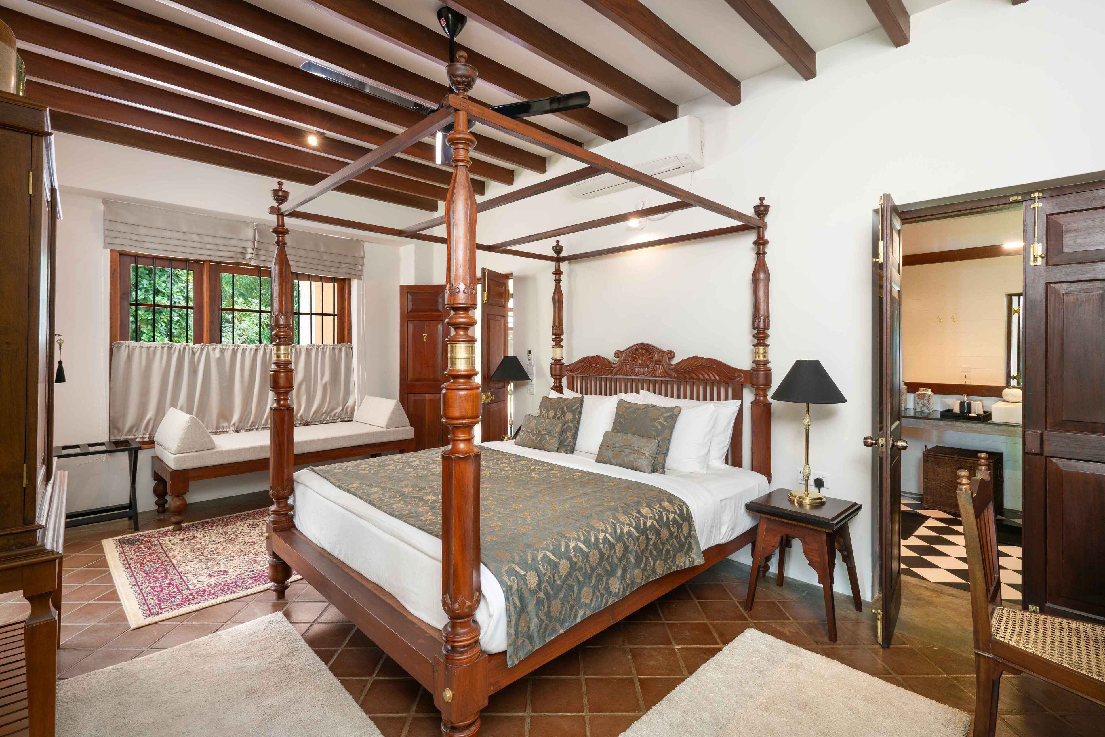 Una cama con dosel de madera y tablero a juego, una mesita de noche con lámpara, y una ventana con cortinas.