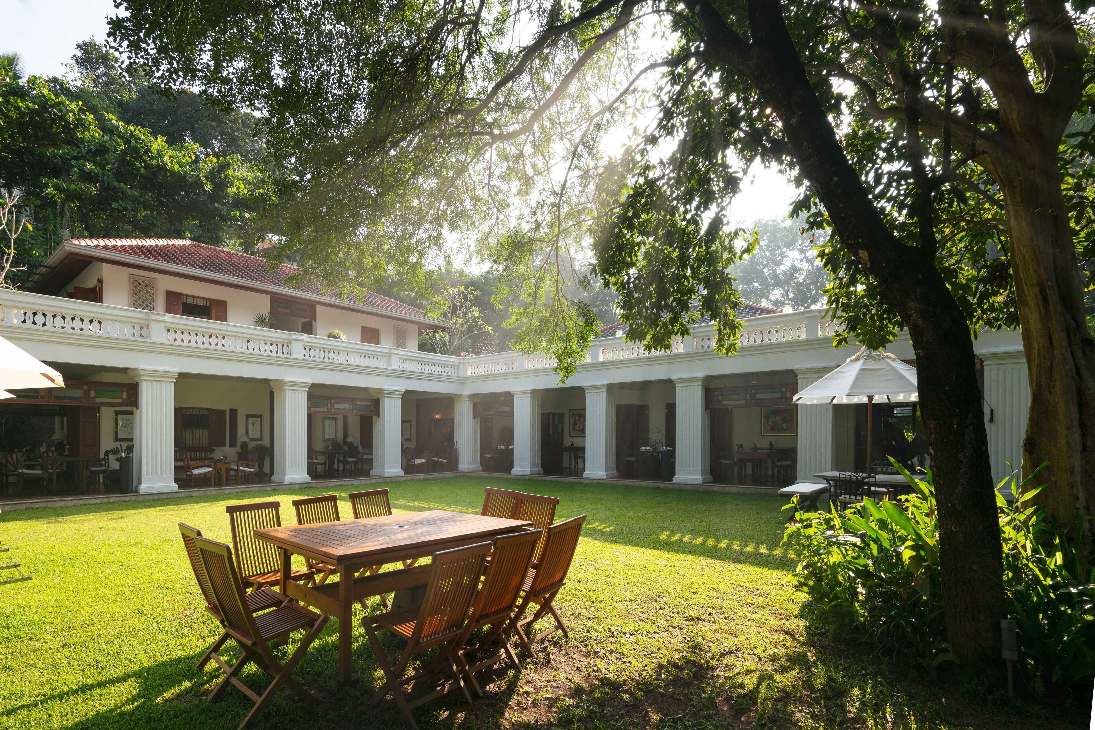 A courtyard with a wooden table and chairs, surrounded by white columns and a building with a red roof.