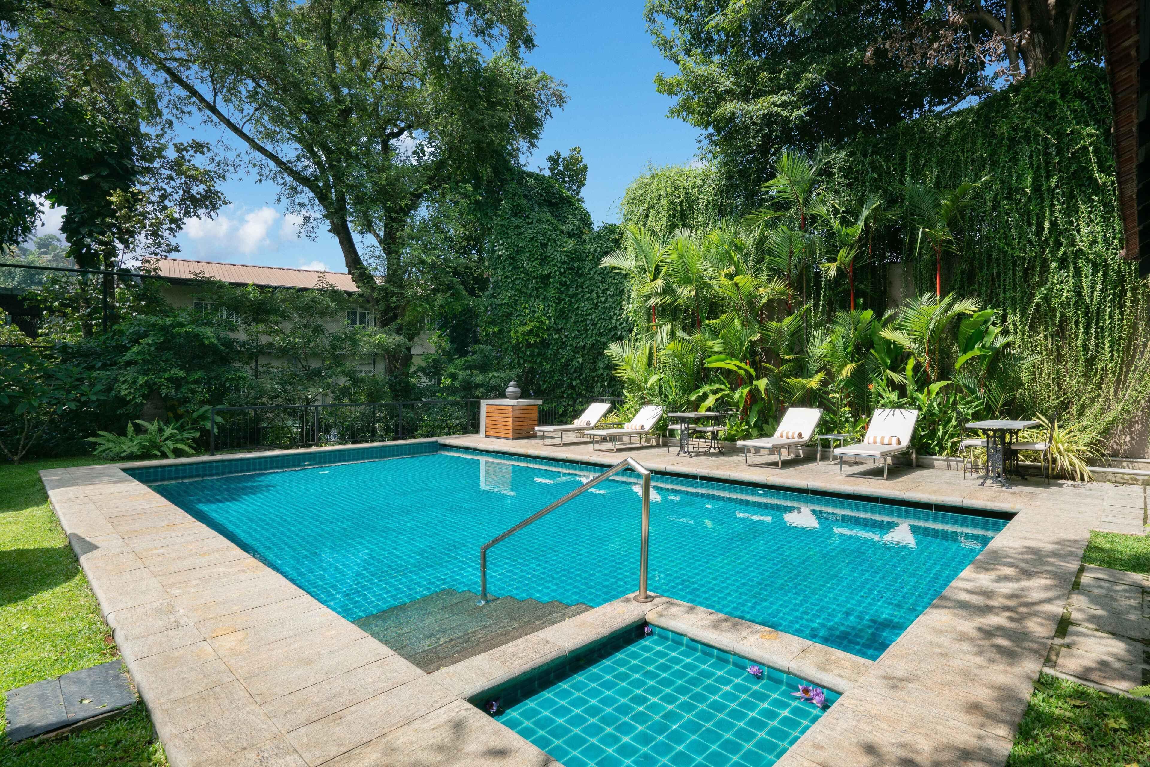 A rectangular swimming pool with a tiled edge, surrounded by a stone deck. There are lounge chairs and a small table with a canopy.