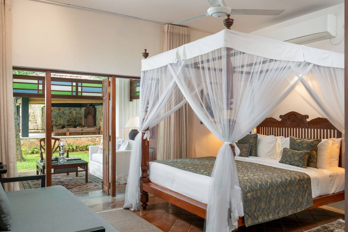 A four-poster canopy bed with a wooden headboard, a nightstand, and a chair in a well-lit room with a view of a garden.