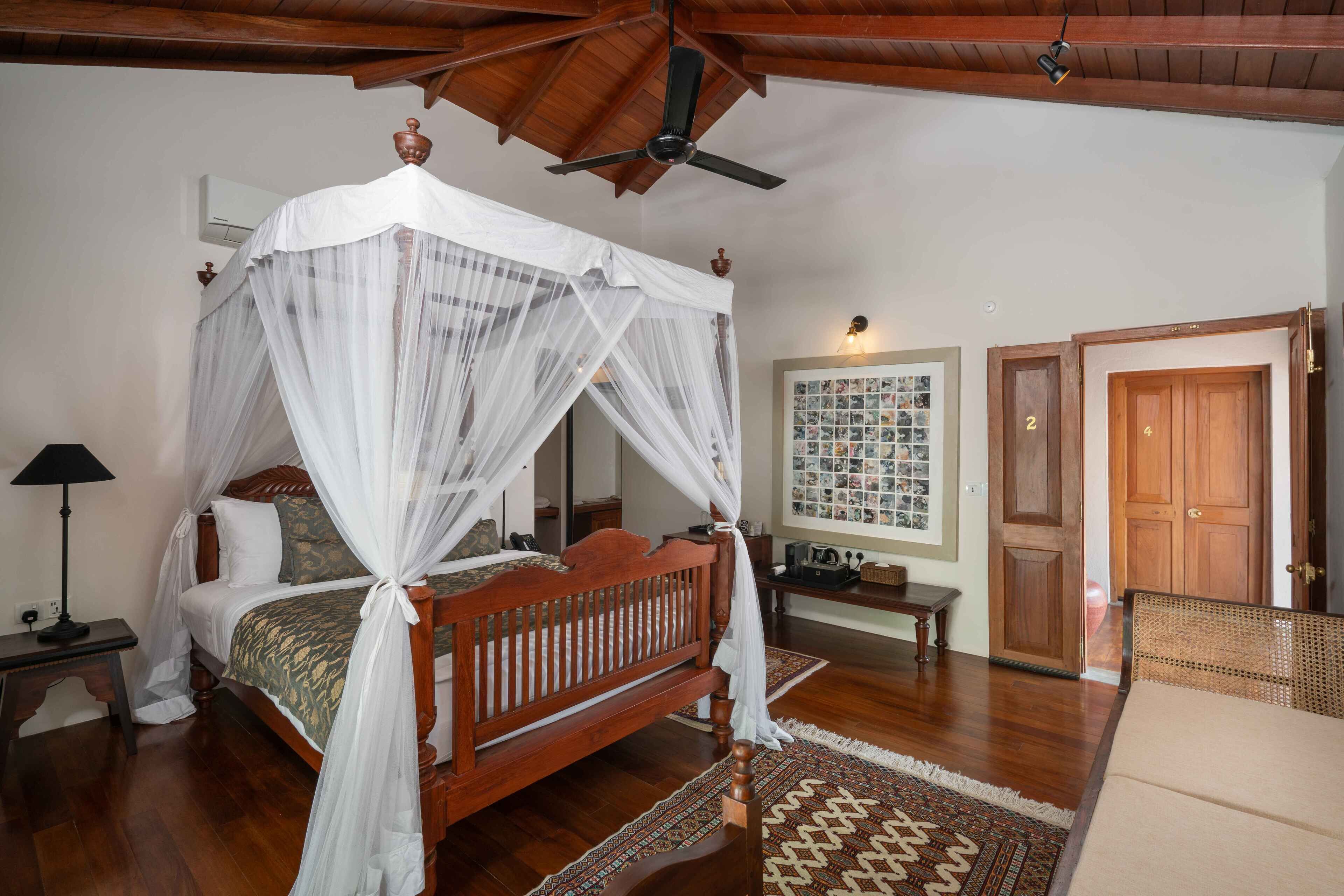 A bedroom with a four-poster bed, wooden ceiling beams, a ceiling fan, a bedside table, a lamp, a wall-mounted air conditioner, and a framed photo collage.