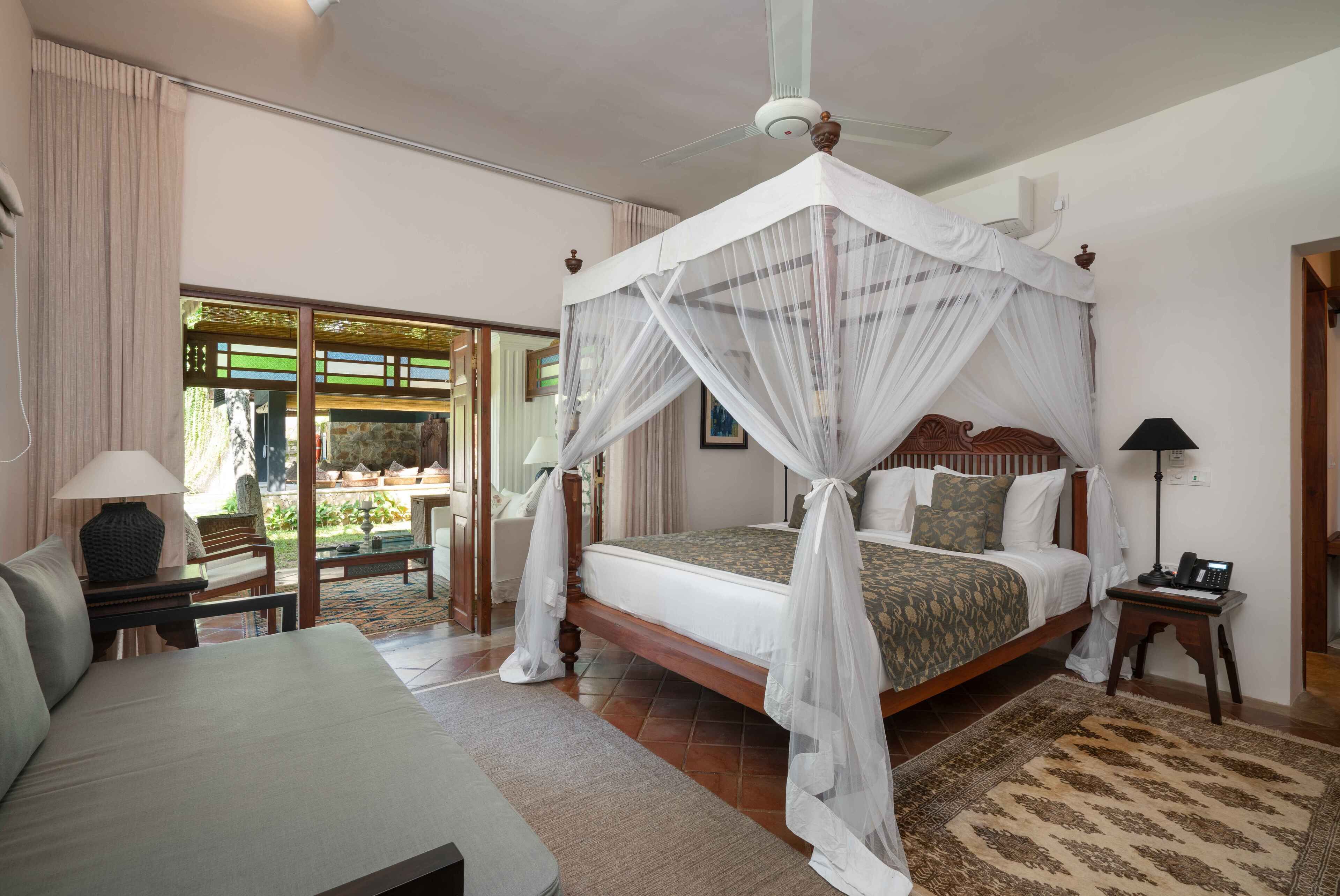 A bedroom with a four-poster bed, a sitting area with a sofa and a table, a bedside lamp, and a view of the outdoors.