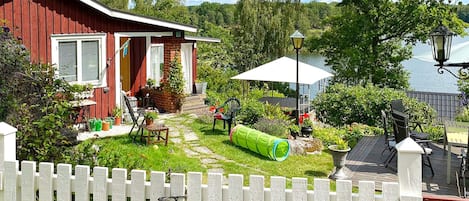 A red house with a white fence in front, a garden with a green tube, and outdoor furniture.