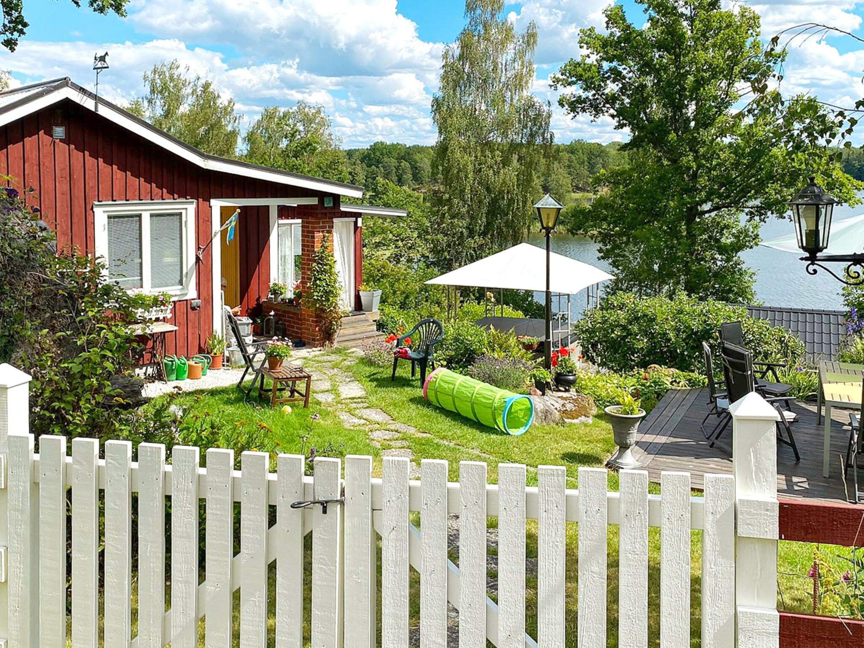 Una casa rossa con una recinzione in legno bianco, un giardino con un tunnel verde e vista su un lago.