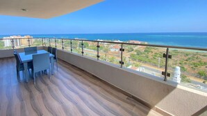 A balcony with a wooden floor, a table, and chairs, overlooking a coastal view with buildings and greenery.