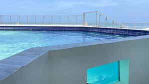 A swimming pool with a clear view of the ocean, a white railing, and a blue-tiled deck.