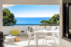 Uma varanda com mesa e cadeiras brancas, uma cesta de vime e vista para o mar.
