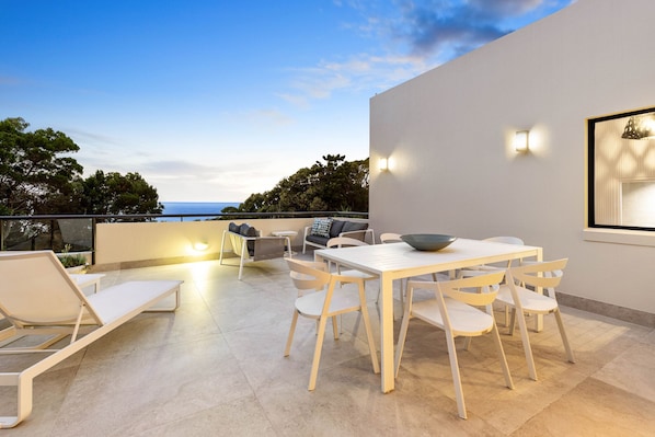 Um pátio moderno ao ar livre com uma mesa e cadeiras brancas, uma poltrona de descanso e vista para o mar.