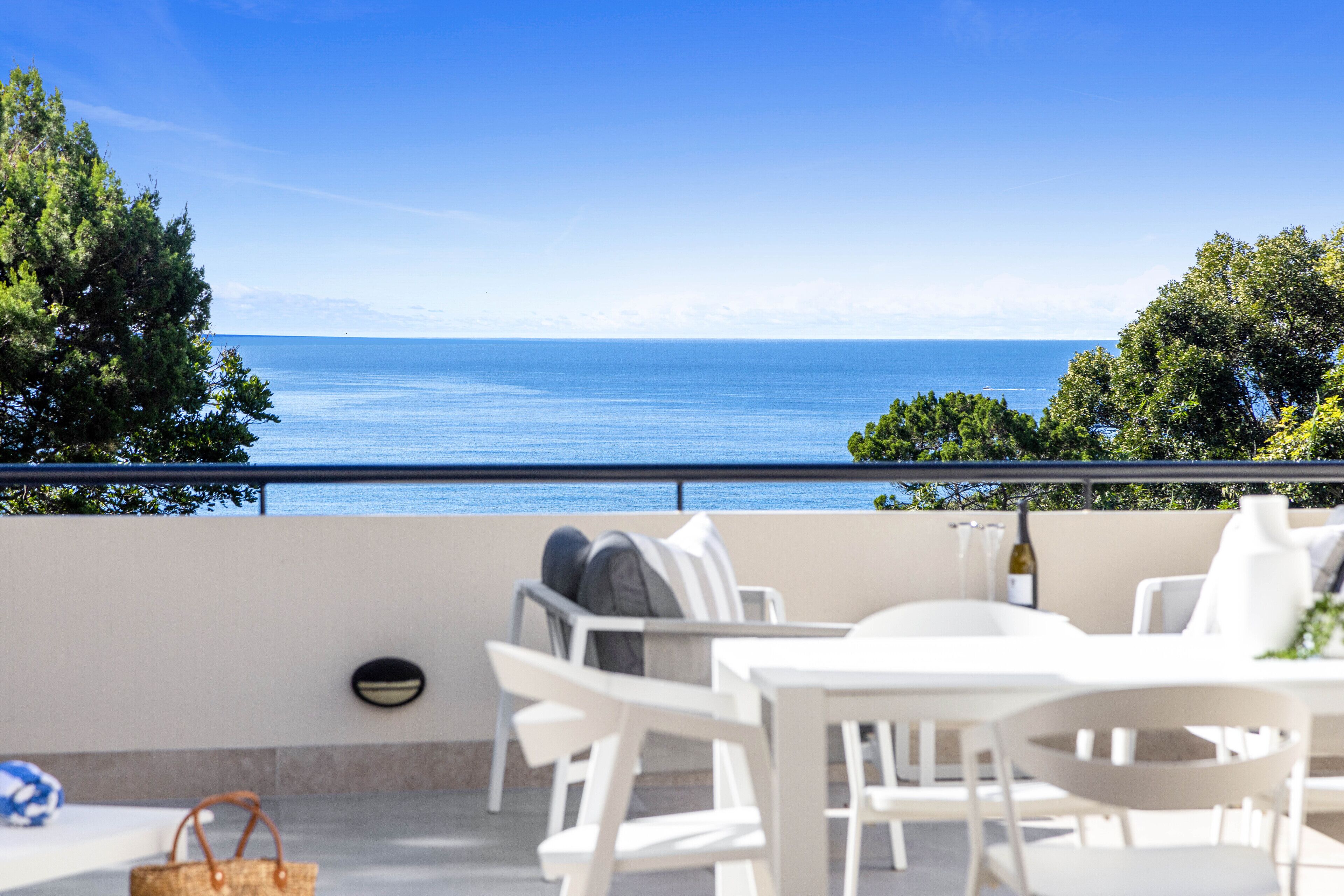 Una terraza con muebles blancos, vista al mar y árboles de fondo.