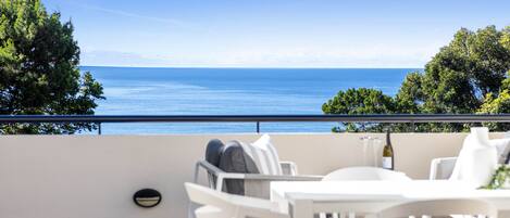 Una terraza con muebles blancos, vista al mar y árboles de fondo.