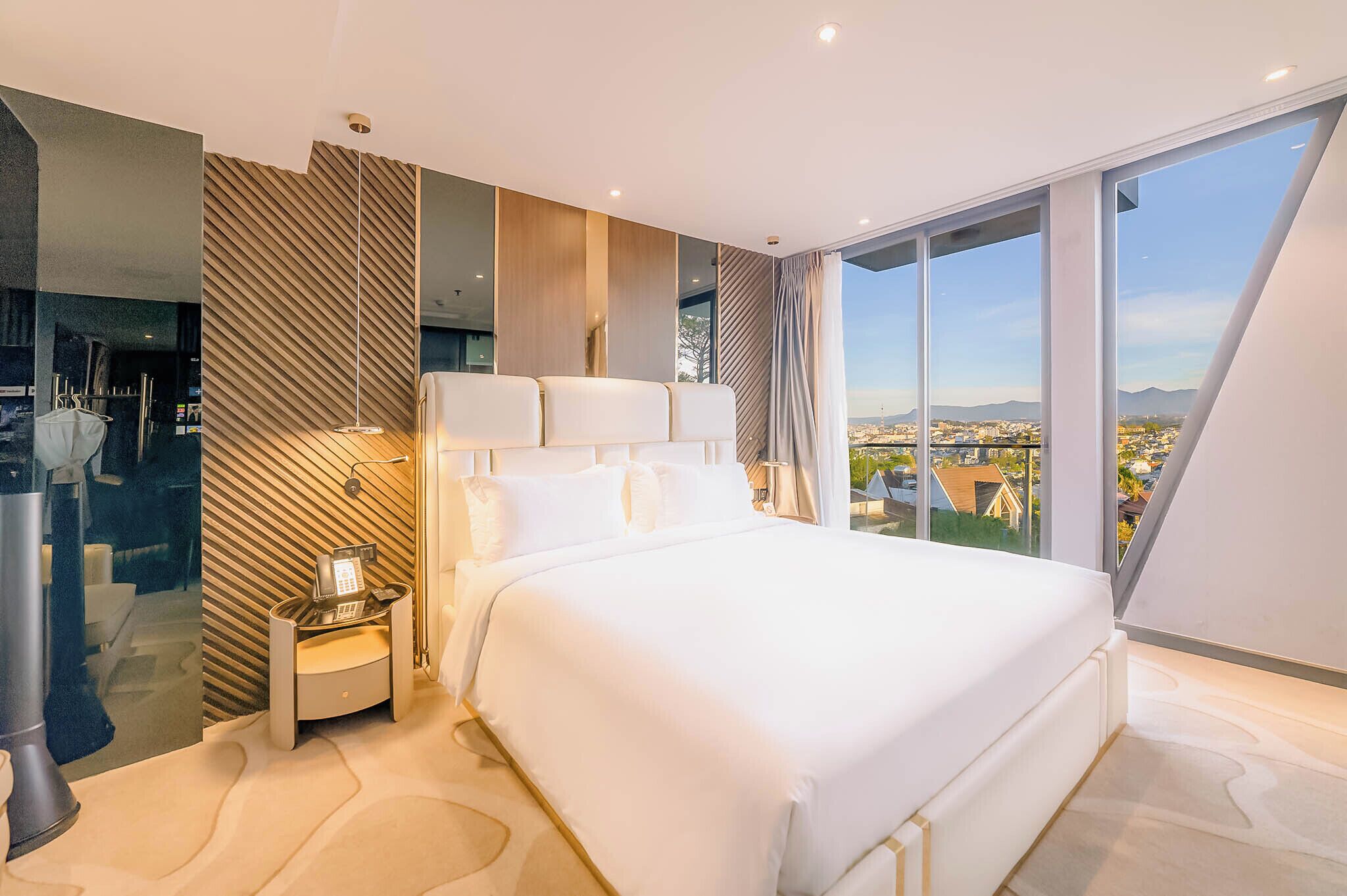 A modern hotel room with a large bed, a bedside telephone, and a city view through large windows.