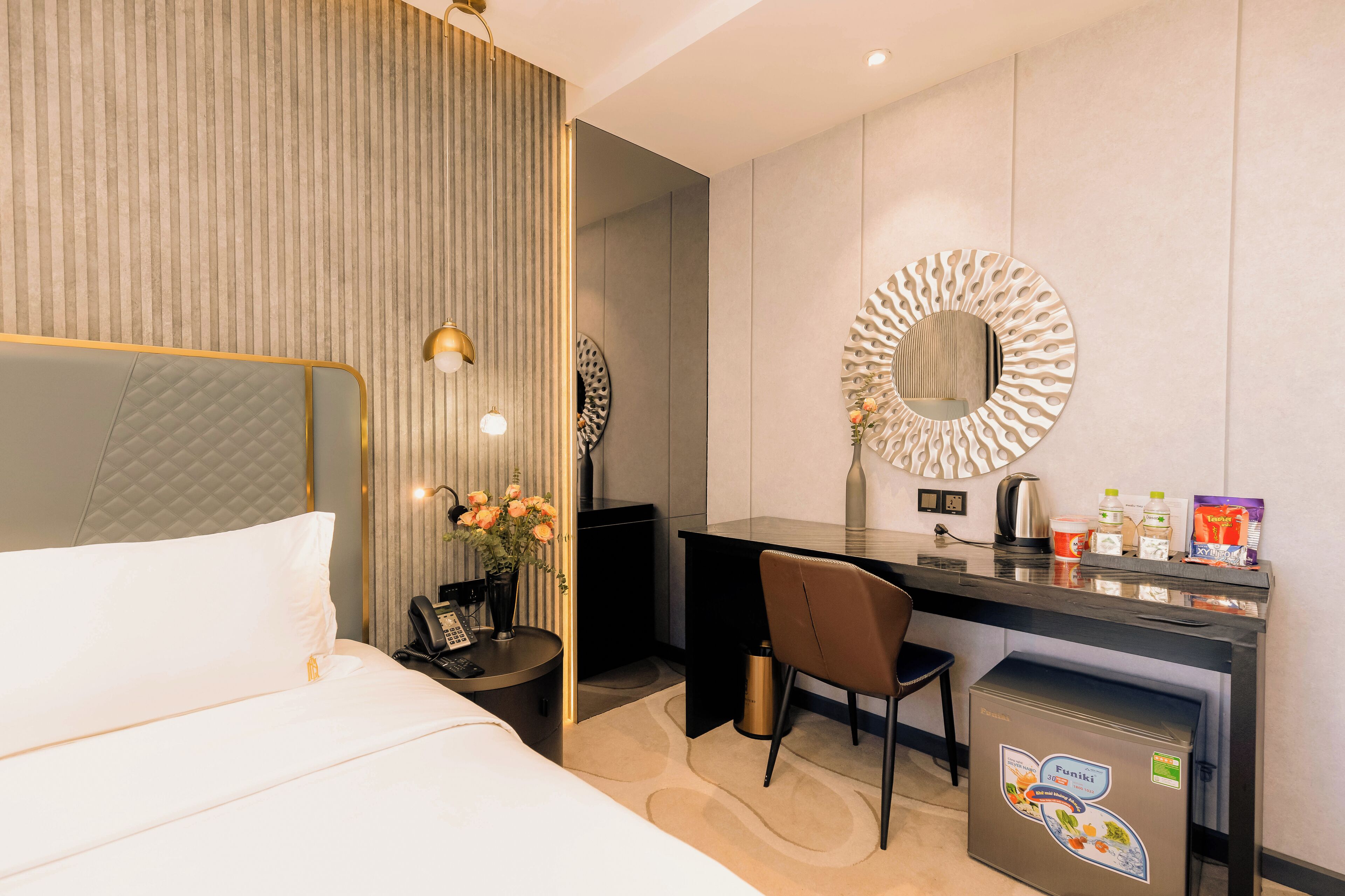 A hotel room with a bed, a desk, a chair, a refrigerator, and a mirror.