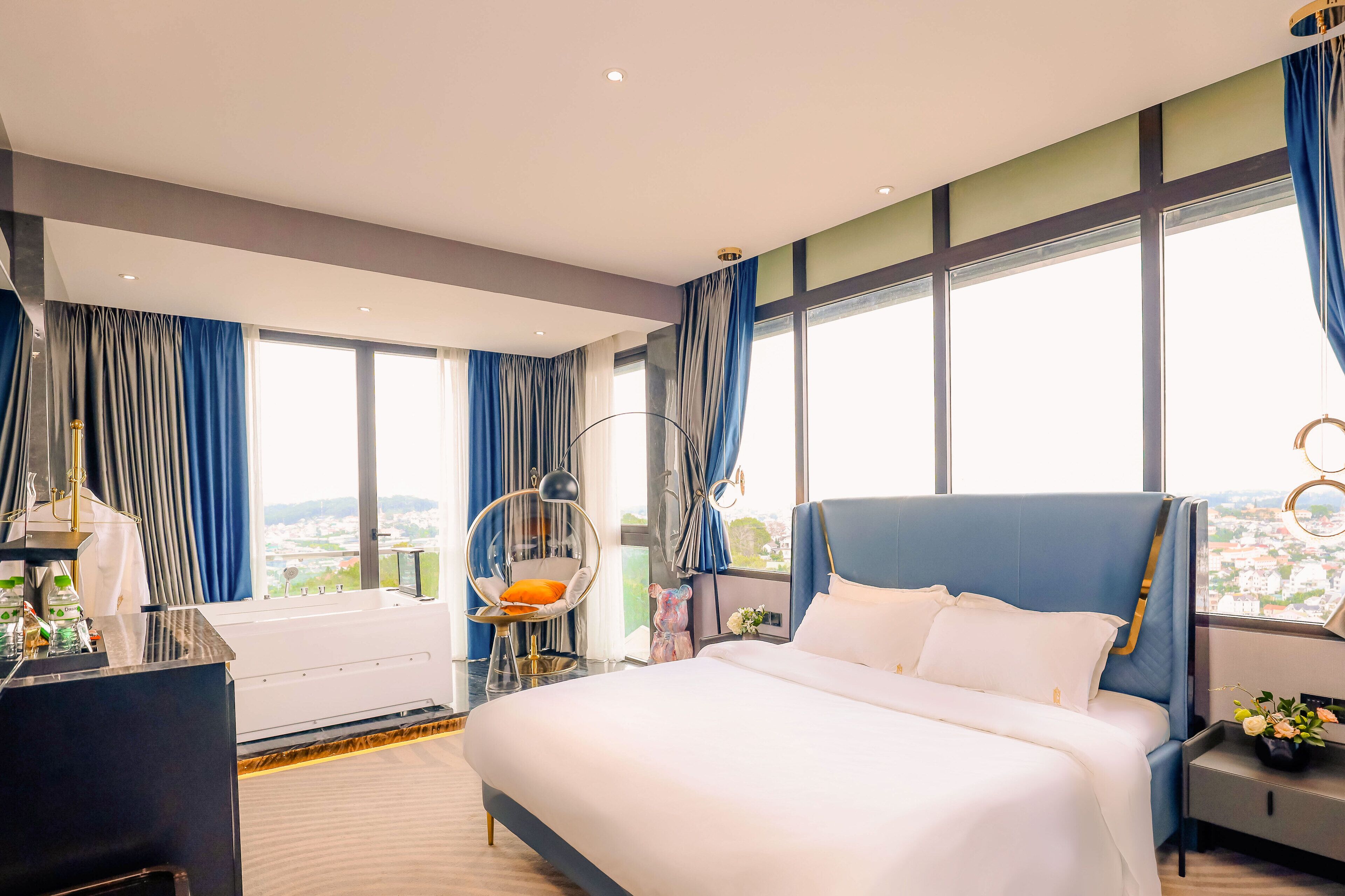 Habitación de hotel moderna con una cama grande, un escritorio y una silla. Amplias ventanas con cortinas, vistas a la ciudad y un elemento decorativo en la pared.