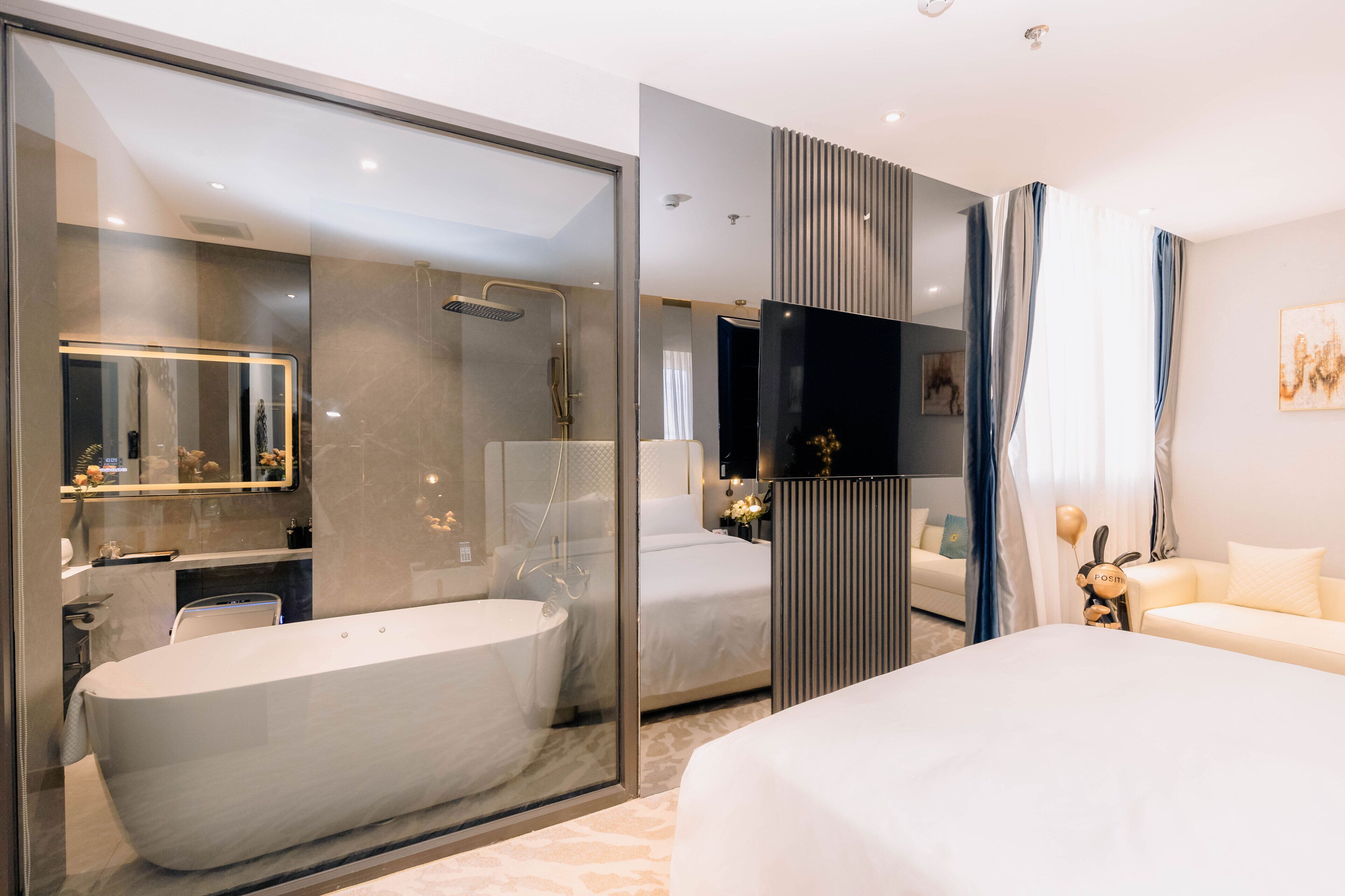 A modern hotel room with a large bathtub, a separate shower area, a flat-screen TV, and a cozy seating area with a sofa and a decorative bunny figure.