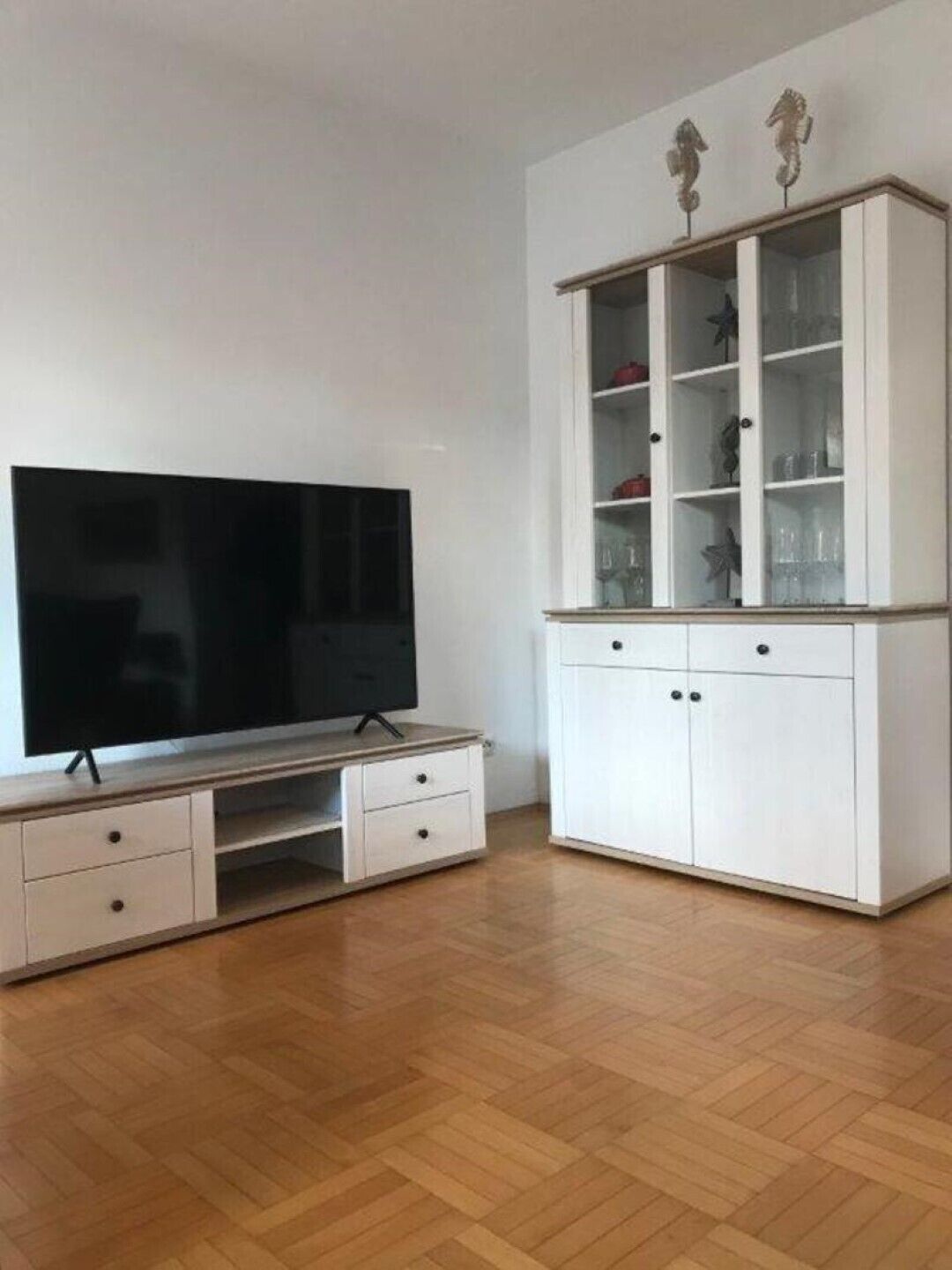 A living room with a flat-screen TV on a wooden TV stand, a white cabinet with glass doors, and a herringbone-patterned wooden floor.