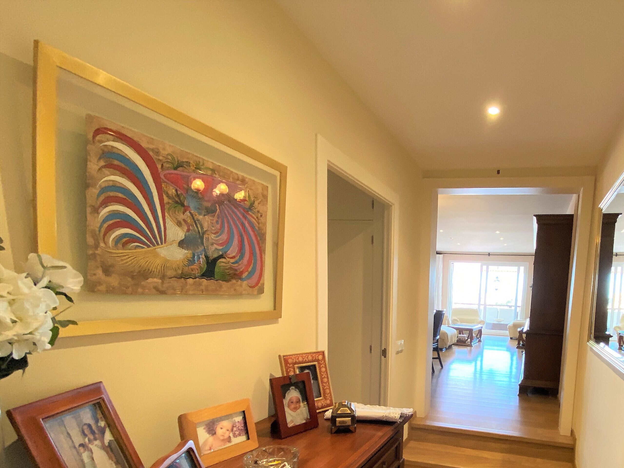 A hallway with a framed artwork, a wooden side table with framed photos, and a vase of white flowers.