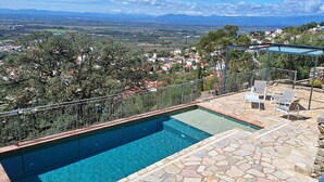 Una piscina en la azotea con solárium de piedra y barandilla de cristal, con vistas a un paisaje pintoresco con casas y árboles.
